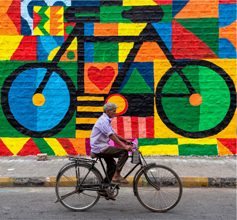 Sanjay Joshi — Jodhpur, India. A cyclist crosses a wall bursting with colour and movement, his quiet journey becoming part of the mural.