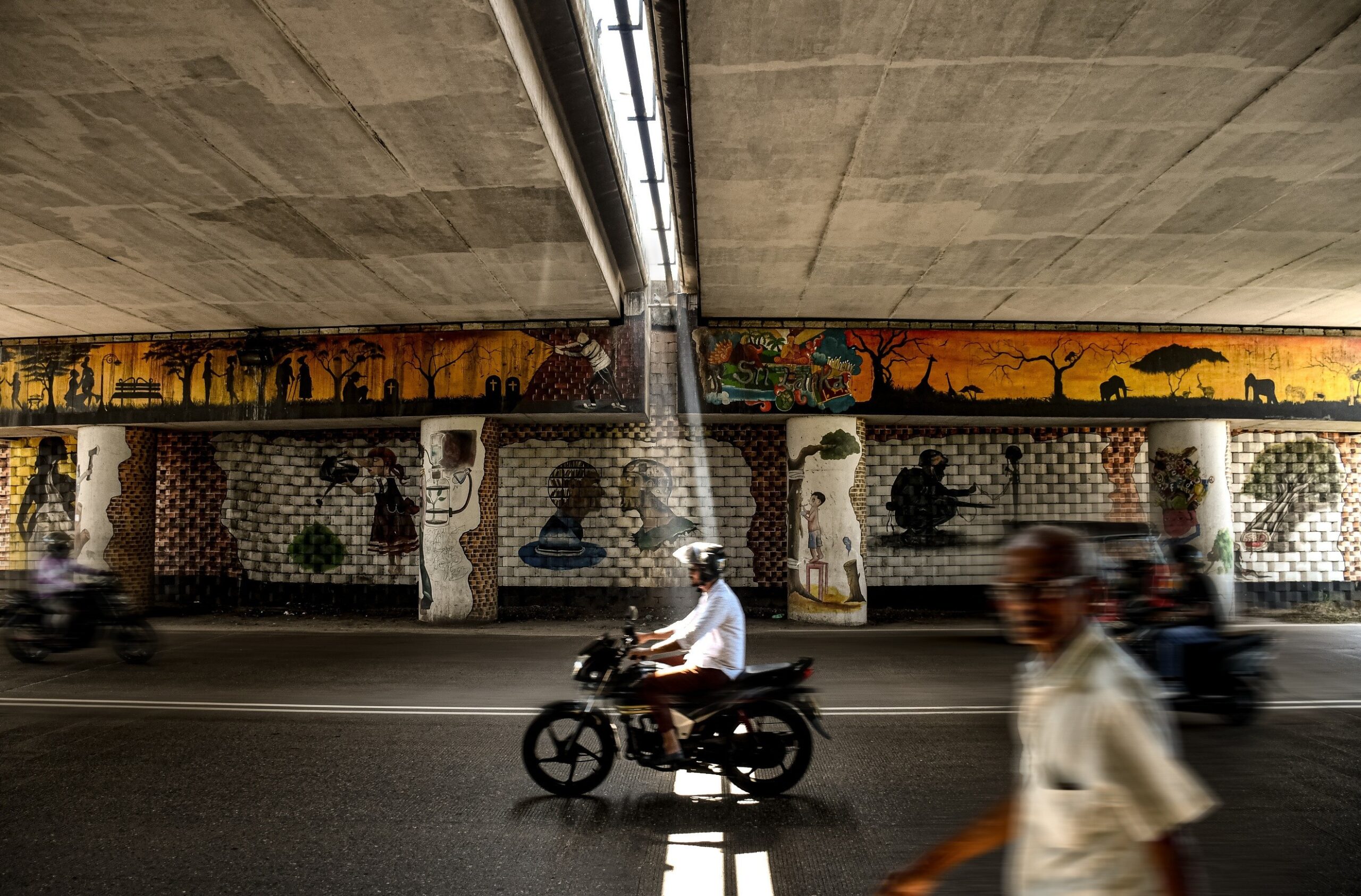 Ranjaka Hettiarachchige — Colombo, Sri Lanka. A single beam of sunlight spotlights a motorcyclist in motion, against a backdrop of hand-painted murals.